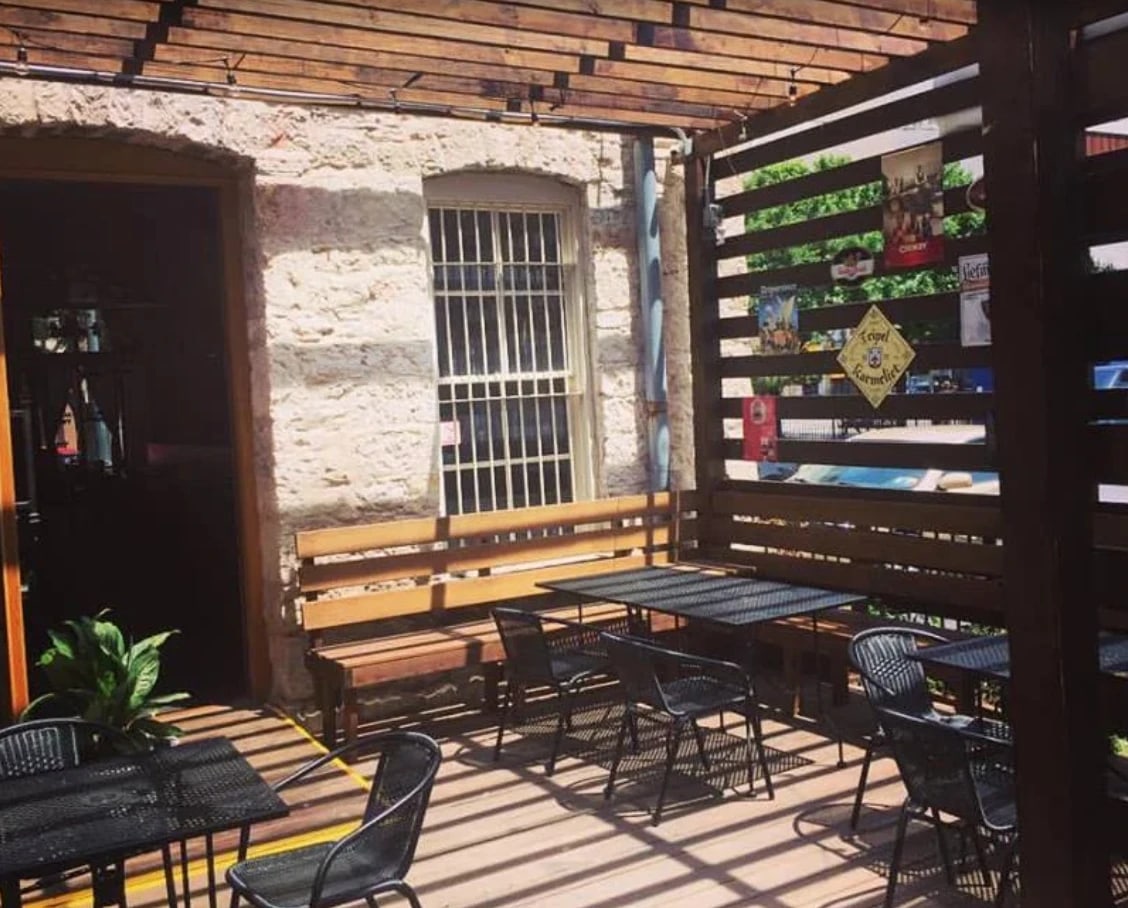 The back courtyard: black metal tables and a wooden bench under a slatted pergola, against the old limestone wall, Belgian beer signs on the wooden screen