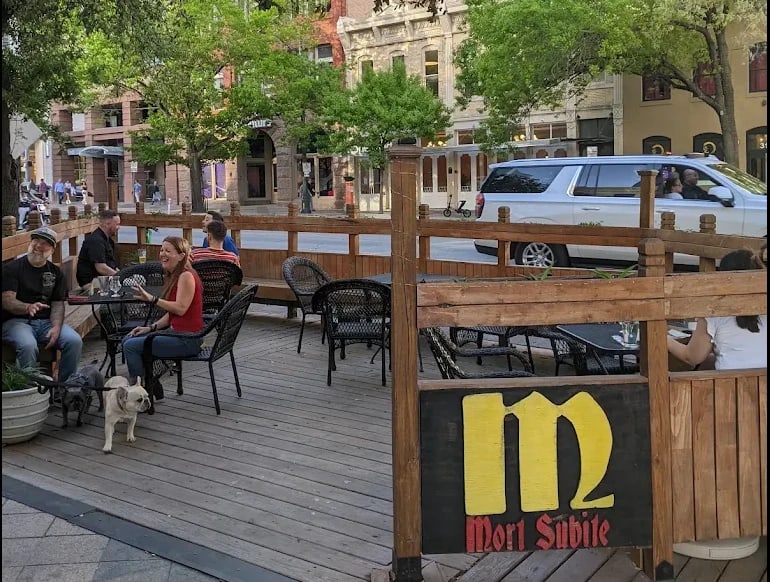The front patio on Congress Avenue: guests at black wicker tables behind a wooden rail, the Mort Subite sign on the rail, the street and trees behind